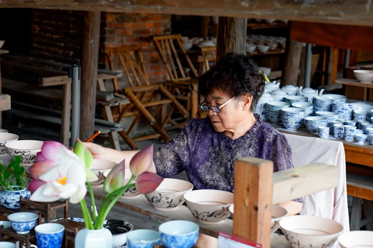 An elderly craftswoman paints a row of unfired bowls with a fine brush, shelves of blue-and-white porcelain behind her