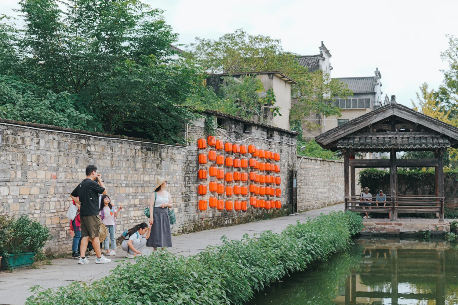 A family walks a stone lane past a wall of red lanterns in Xixinan village while an older man rests in the covered bridge pavilion by the water