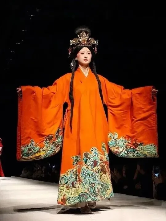 Performer in an orange wide-sleeved robe with an embroidered wave hem, arms outstretched