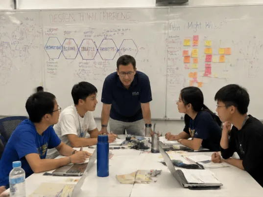 A mentor in a UCLA shirt guiding five students at a table beneath a design-thinking whiteboard.