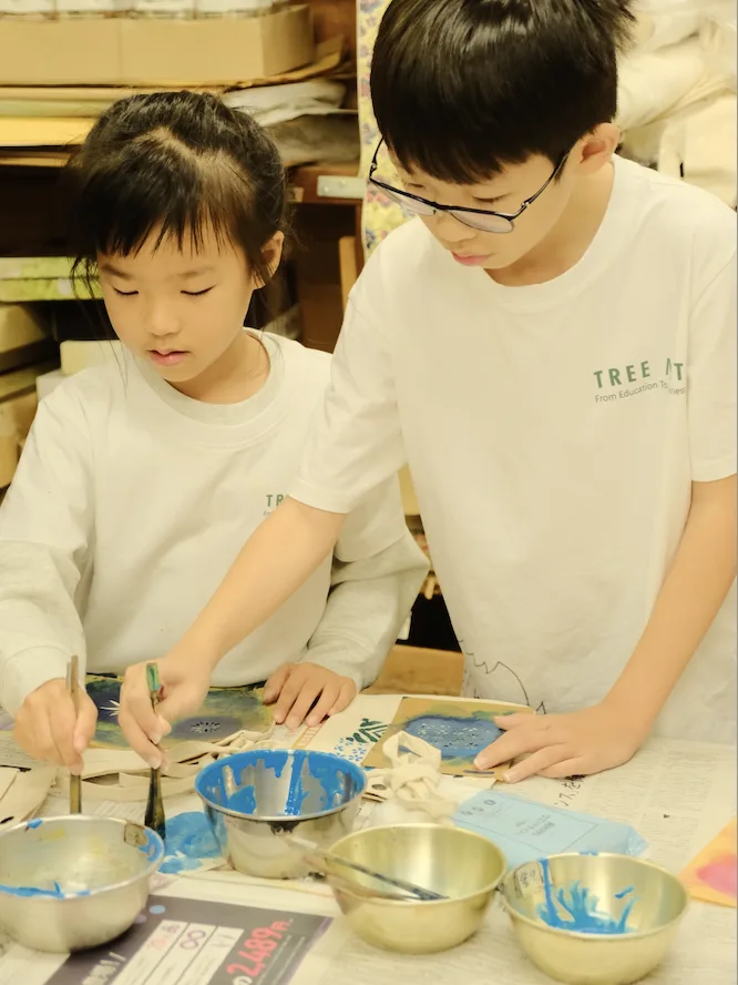 Two children brushing blue dye onto stencilled cloth beside bowls of pigment
