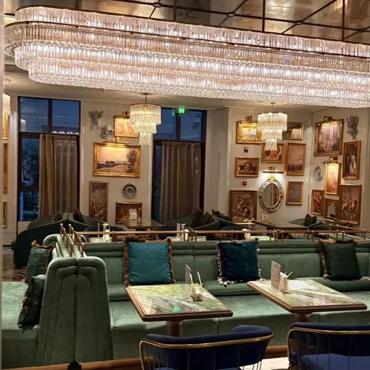 A hotel lounge with a long crystal chandelier, green velvet banquettes and gilt-framed paintings