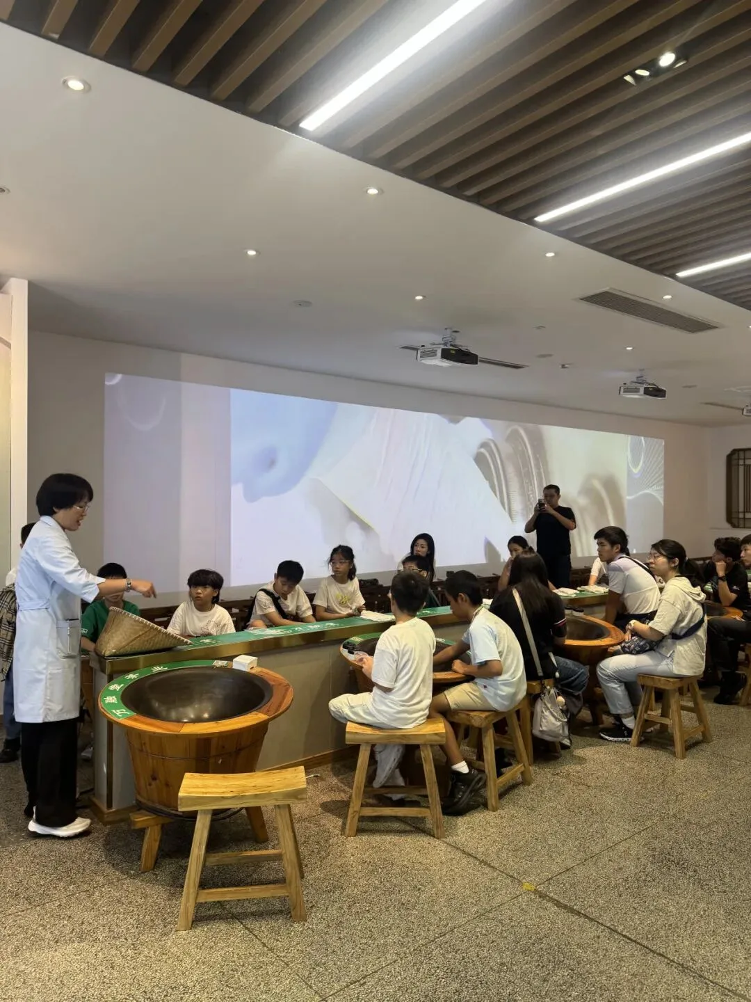A class works at a row of tea-roasting woks in front of a projected film
