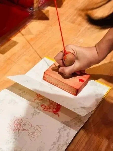 Hand pressing a large red-inked seal block onto paper over a printed bodhisattva line drawing