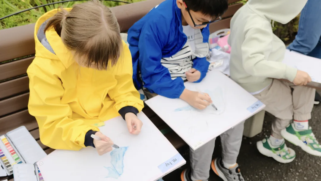 Three children painting Mount Fuji in watercolour on boards resting on a bench