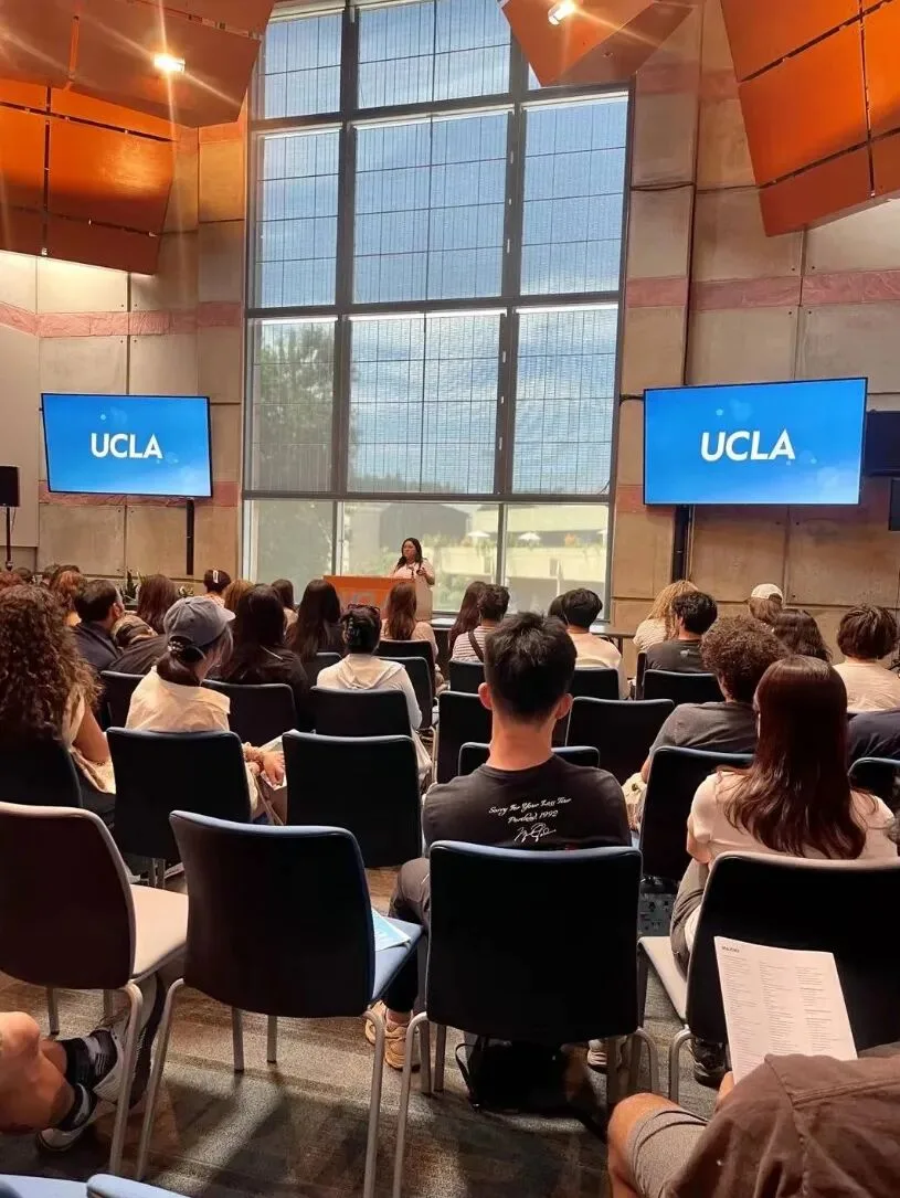 A speaker addressing a seated audience between two UCLA screens in a tall glass-walled hall.