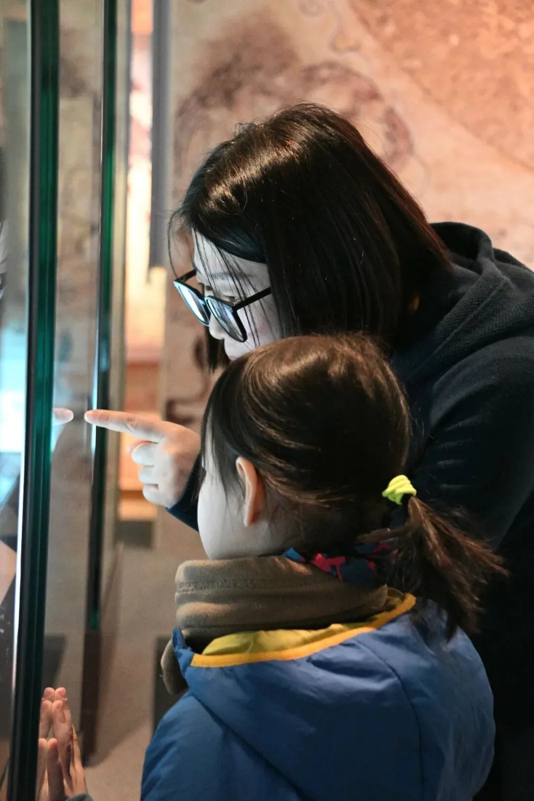 Woman pointing into a museum display case for a girl, a mural-painted wall behind