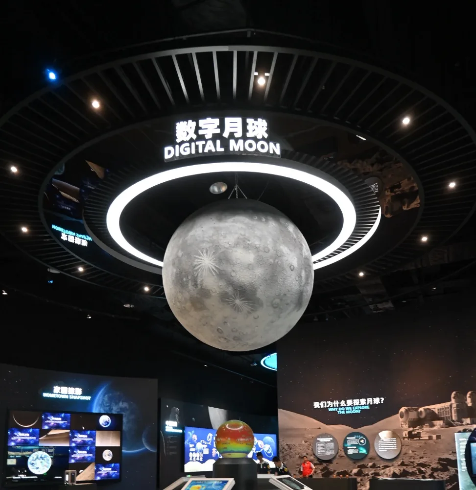 A lit lunar globe hangs beneath a ring-shaped Digital Moon display in a dark exhibition hall.