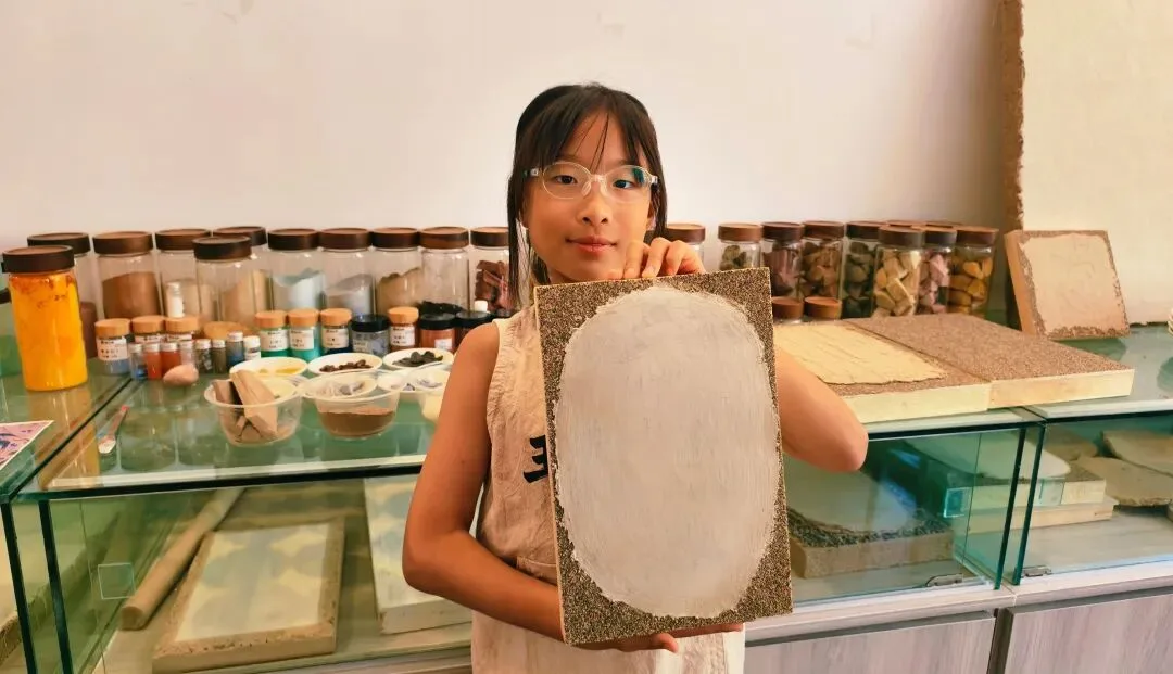 Girl holding up a clay board primed with a white oval, shelves of pigment jars behind