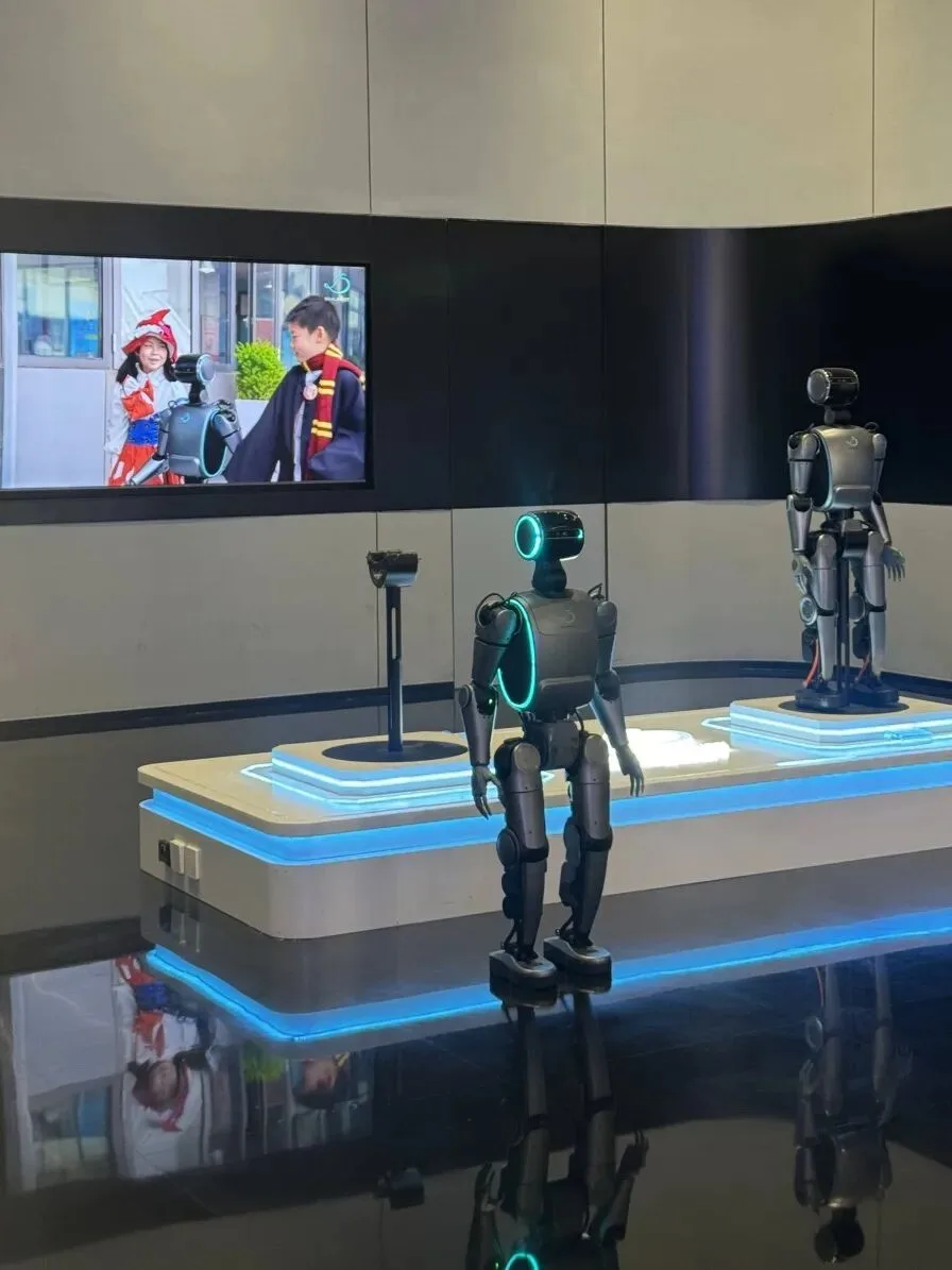 Two humanoid robots stand on a lit display plinth in front of a video screen