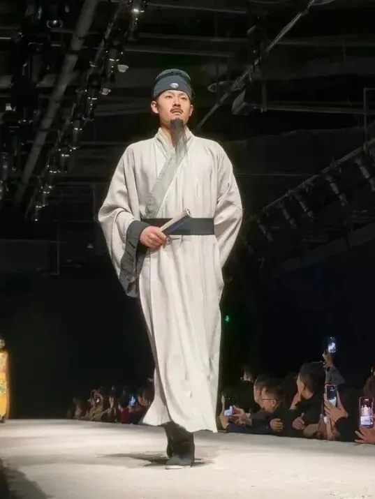 Performer in a pale scholar's robe and headwrap walking a runway before a seated audience