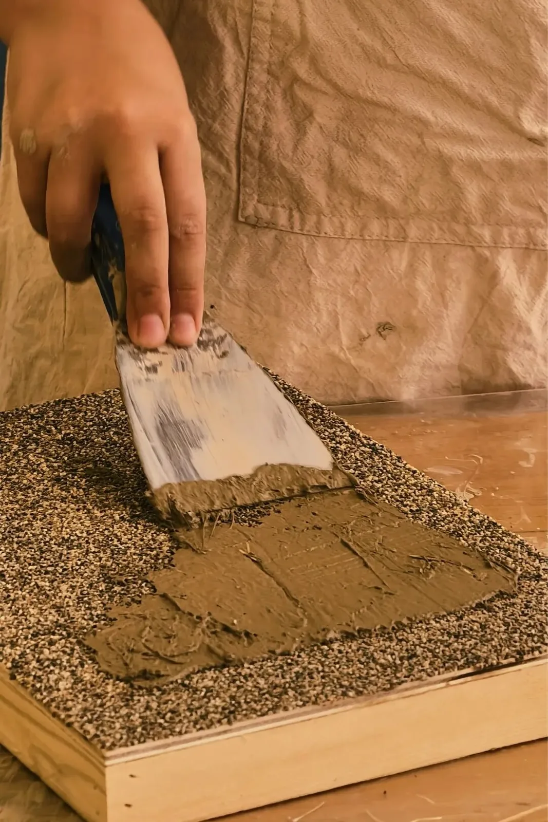 Brown clay spread with a metal spatula across a grit-covered wooden mural board