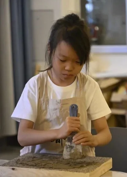 Girl in an apron spreading grey clay across a grit-edged board with a metal spatula