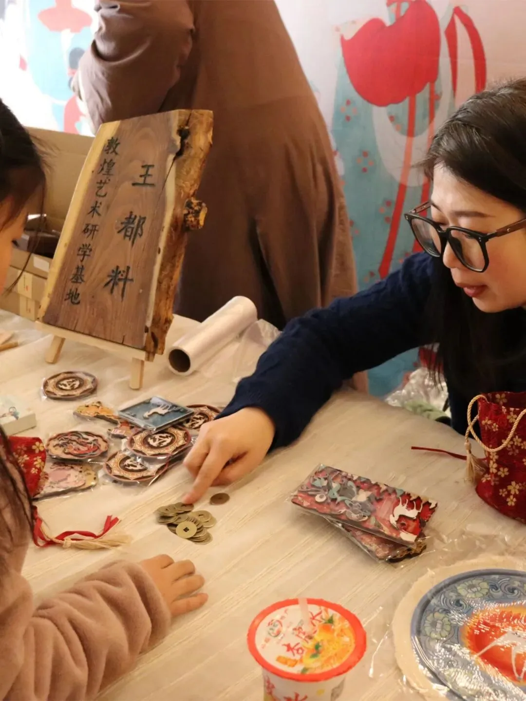 Stallholder and children over a table of Dunhuang souvenir badges and a pile of bronze coins