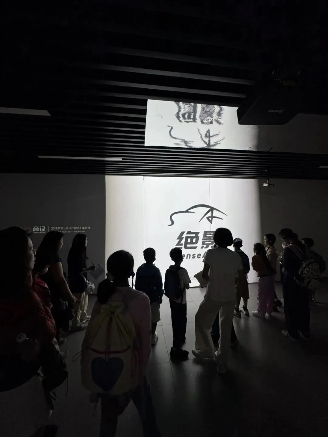 Children stand in a darkened hall watching a projected car logo on the wall