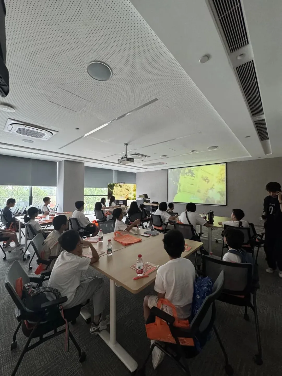 Children sit at long meeting-room tables watching a projected presentation