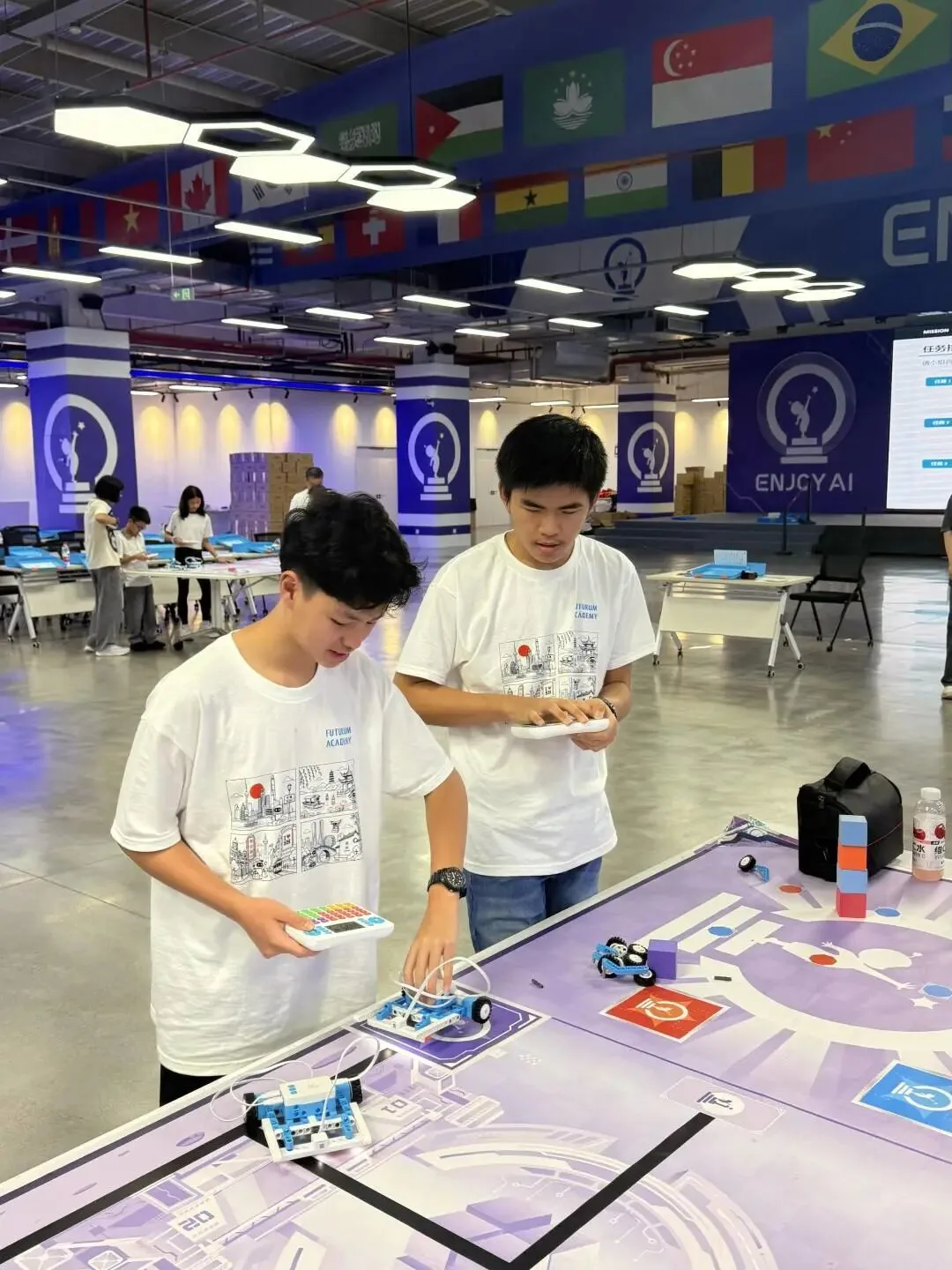 Two teenagers with handheld controllers drive small robot cars across a competition mat