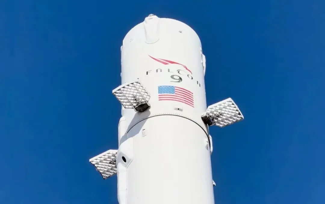 The top of a Falcon 9 booster, grid fins folded against the fuselage, under a clear sky.