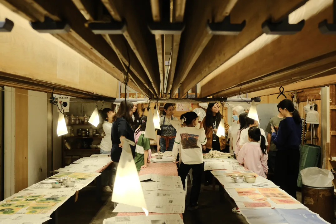 Students walking through a dyeing workshop lined with long tables of drying dyed panels