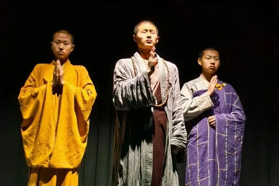 Three performers in yellow, grey and purple monk robes, palms pressed together on a dark stage