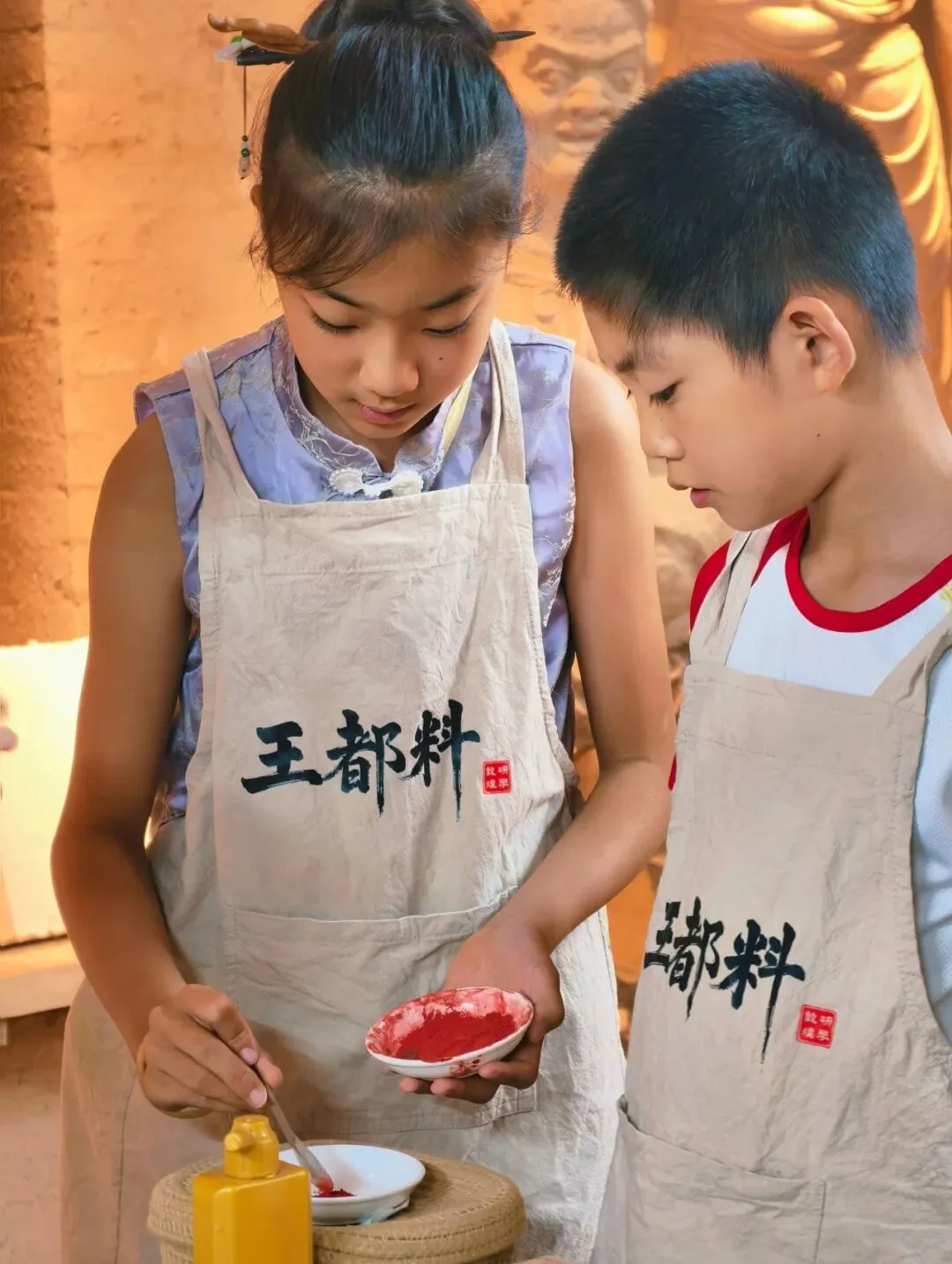 Girl stirring red pigment in a saucer while a boy watches, a gilded statue behind