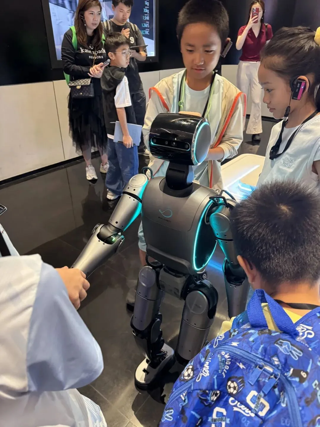 Children gather around a teal-lit humanoid robot, one of them shaking its mechanical hand