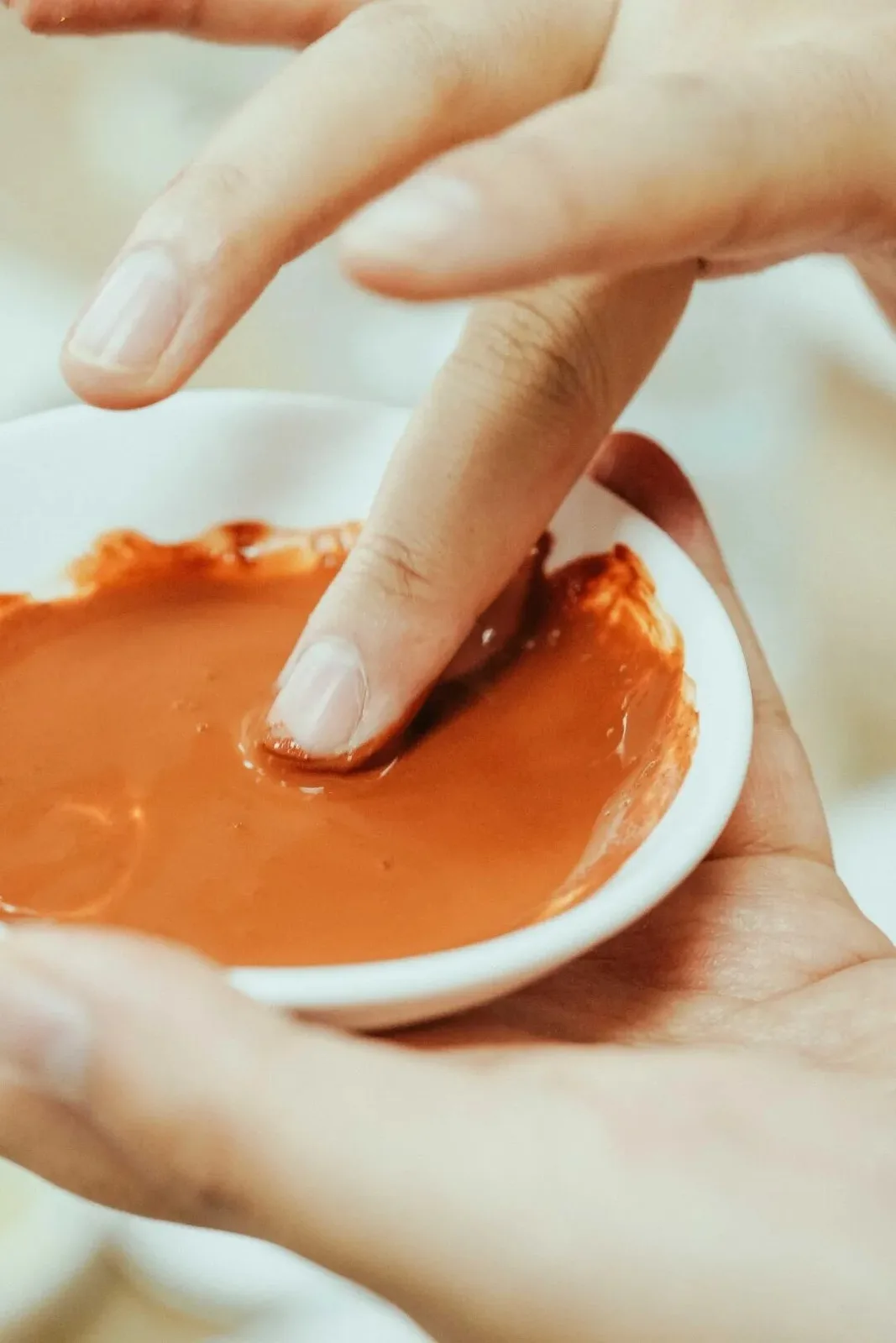 A fingertip testing a bowl of orange-red pigment mixed with glue