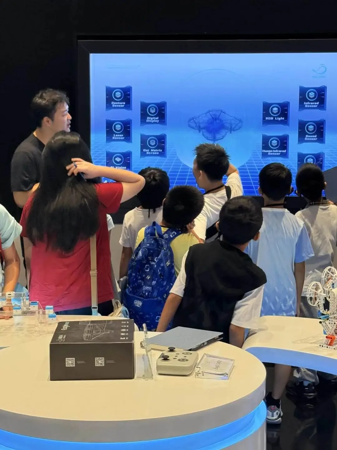 A guide explains a drone to children in front of a screen labelling gesture, laser and infrared sensors