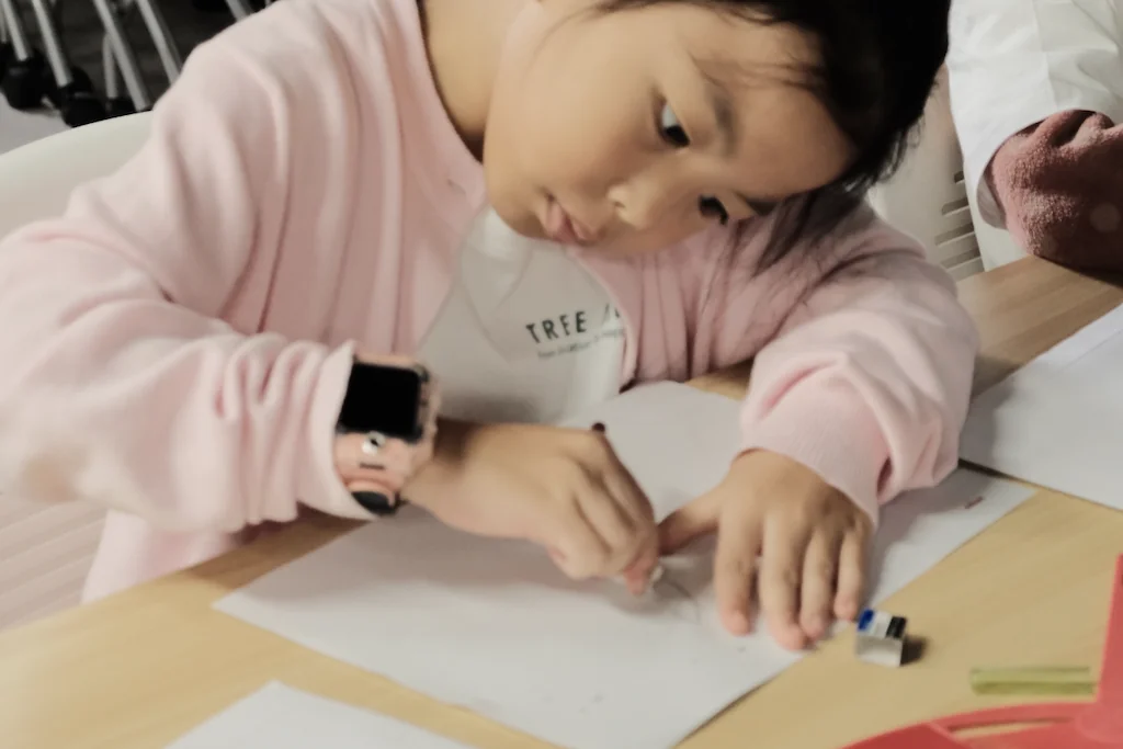 A girl in a pink hoodie and TREE ART apron leaning close over her drawing