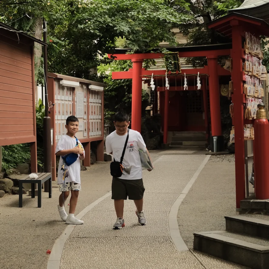Two boys walking past a red torii gate and a rack of wooden ema plaques