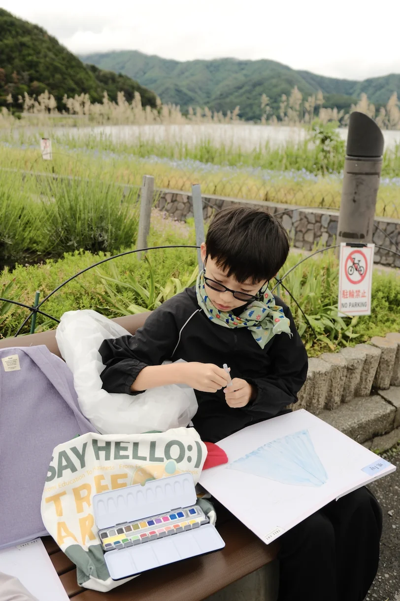A boy in glasses painting a blue Mount Fuji in watercolour beside the lake