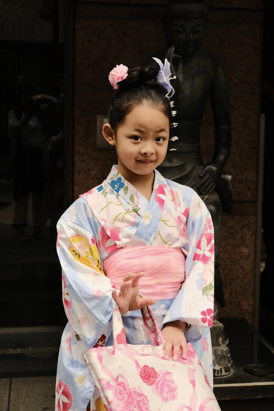 A girl in a pale blue floral kimono and pink obi in front of a bronze Buddha statue